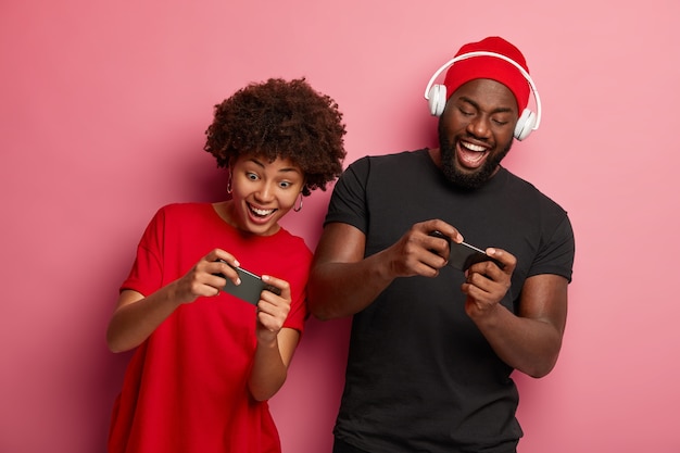 Happy Afro American girlfriend and boyfriend play games on cellphones, compets in online competition, have fun together