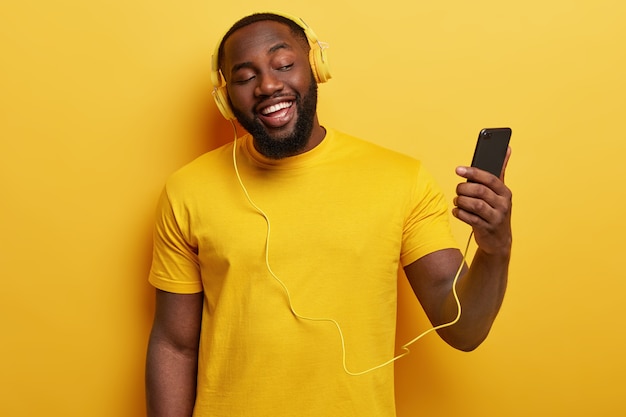 행복한 아프리카 계 미국인 남자가 함께 노래하는 노래를 즐기고 헤드폰에 연결된 현대 휴대 전화를 보유하고 있습니다.