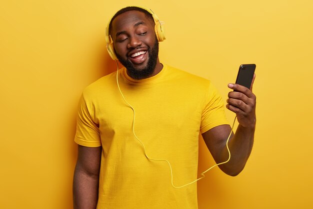 Happy Afro American man enjoys song to sing along, holds modern cell phone connected to headphones