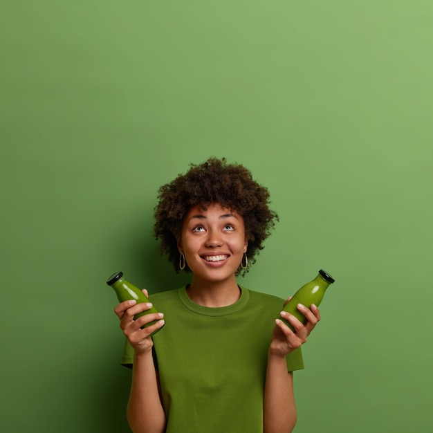 무료 사진 행복한 아프리카 계 미국인 여성은 차가운 건강 혼합 음료를 들고 위에 집중하고 녹색 비건 야채 스무디를 마시고 건강한 상태에 집중하고 즐겁게 미소 짓습니다. superfood 개념