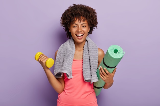 Free photo happy afro american woman lifts small weights, holds green karemat, ready for yoga exercises