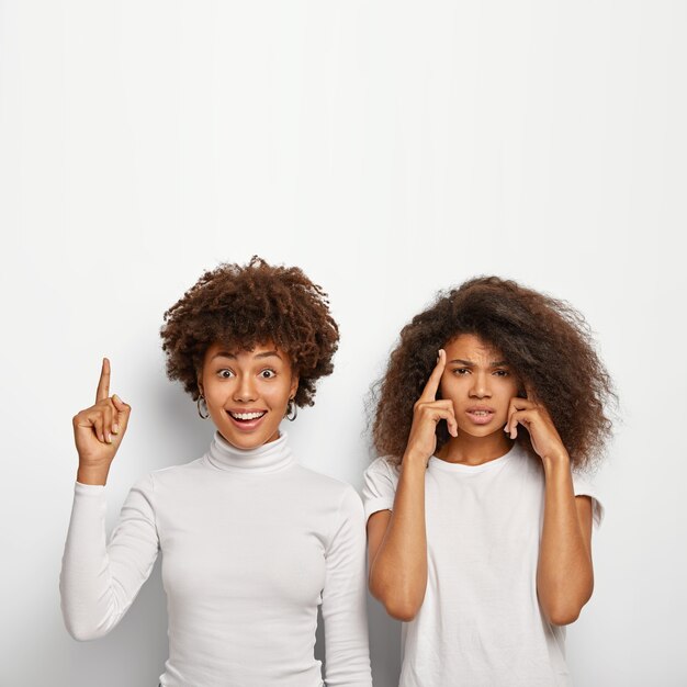 Happy Afro American woman points above fore finger, her intense friend touches temples, tries to recall important information