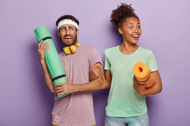 Free photo happy afro woman with fitness mat, holds mans hand, asks to go with her on training, displeased man has no desire for sport workout, wears headband, t shirt and headphones. couple with mats