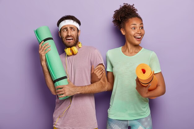 Happy Afro woman with fitness mat, holds mans hand, asks to go with her on training, displeased man has no desire for sport workout, wears headband, t shirt and headphones. Couple with mats