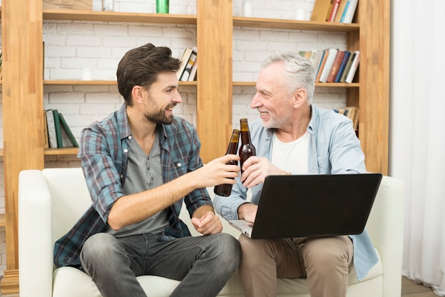 Happy aged man and young guy clanging bottles and using laptop on sofa