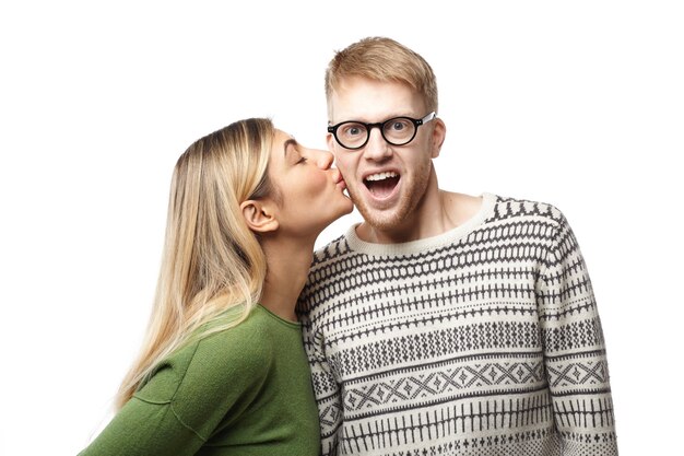 happy amazed young geeky guy with beard wearing eyeglasses and sweater opening mouth excitedly while being kissed by attractive blonde woman on cheek. Love and romance concept