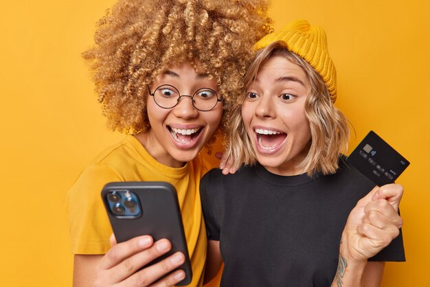 幸せな驚きの若い女性は、オンラインショッピングで携帯電話とクレジットカードのダウンロードアプリケーションを使用して、黄色の背景にさりげなく隔離された服を着た最新のテクノロジーを使用して支払いを行います