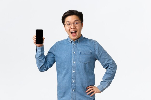 Счастливый и удивленный красивый азиатский парень с подтяжками и очками реагирует на фантастические новости, показывает экран мобильного телефона, представляет приложение или магазин, стоя на белом фоне изумленно.