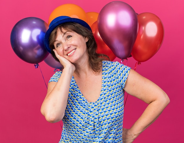 Счастливая и веселая женщина среднего возраста в партийной шляпе, держащая разноцветные воздушные шары, глядя вверх с улыбкой на лице, празднует день рождения, стоя над розовой стеной