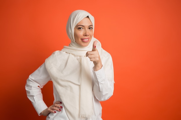 Hijab에서 행복 한 아랍 여자입니다. 빨간 스튜디오 배경에서 카메라를 가리키는 웃는 소녀의 초상화. 젊은 감정적 인 여자. 인간의 감정, 표정 개념.