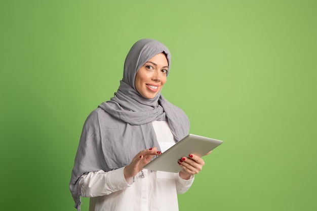 노트북과 hijab에서 행복 한 아랍 여자입니다. 녹색 스튜디오 배경에서 포즈 웃는 소녀의 초상화. 젊은 감정적 인 여자. 인간의 감정, 표정 개념. 전면보기.