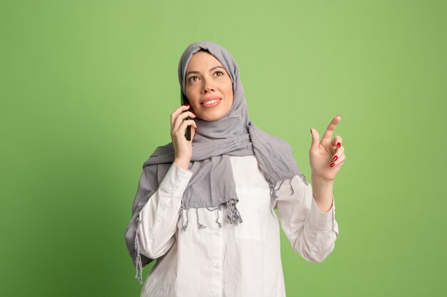 Happy arab woman in hijab with mobile phone. Portrait of smiling girl, posing at green studio background. Young emotional woman. human emotions, facial expression concept. Front view.