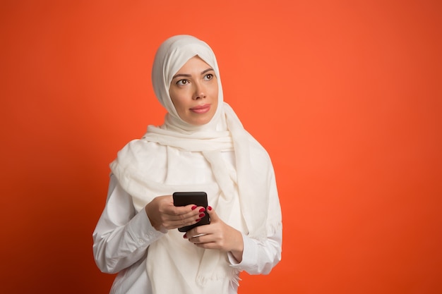 Happy arab woman in hijab with mobile phone. Portrait of smiling girl, posing at red studio background. Young emotional woman. The human emotions, facial expression concept. Front view.