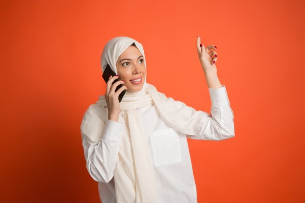 휴대 전화와 함께 hijab에서 행복 한 아랍 여자입니다. 빨간 스튜디오 배경에서 포즈 웃는 소녀의 초상화.