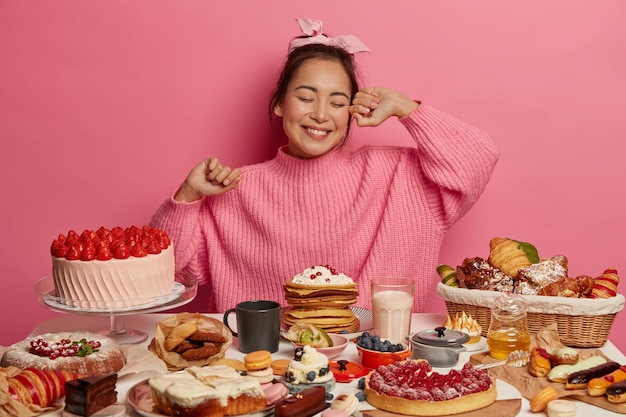 La ragazza asiatica felice di compleanno arriva al tea party, mangia dolci deliziose torte, circondata da molti dessert, pone su sfondo rosa.