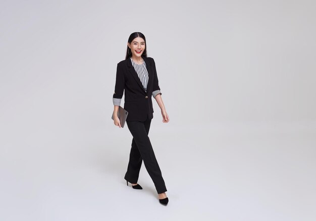 태블릿을 들고 회색 배경 위를 걷는 정장을 입은 행복한 아시아 비즈니스 여성 미소