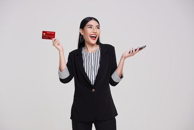 携帯電話を使用して、灰色の背景に分離されたクレジット カードを示す幸せなアジア ビジネス女性