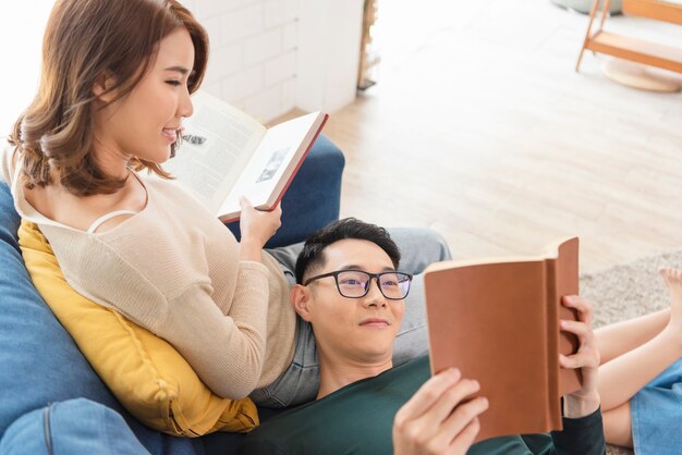 Happy asian couple is spending weekend together on couch indoors at home, relaxing and enjoying reading book.