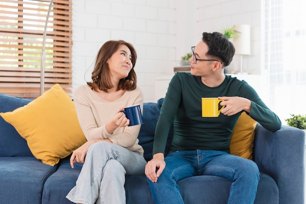 Happy Asian couple man and woman is spending weekend together on couch indoors at home, relaxing and enjoying drinking coffee.