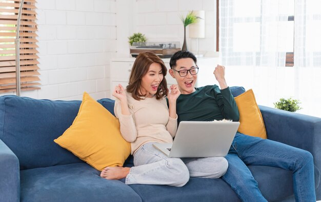 Happy Asian couple man and woman is spending weekend together watching movie on couch indoors at home, relaxing and enjoying eating popcorn.