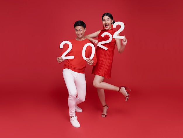 Happy asian couple in red casual attire showing number 2022 greeting happy new year with smiles on bright red background.