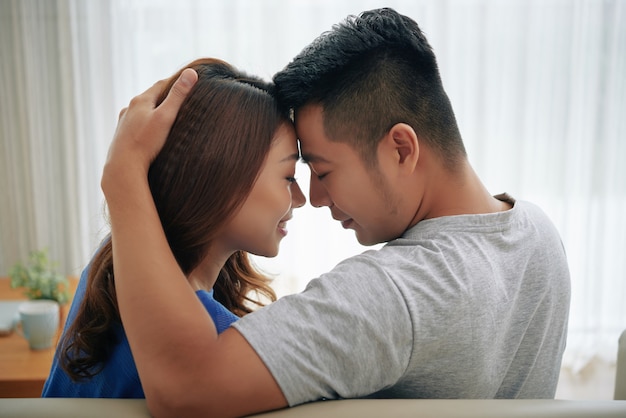 Happy Asian couple sitting on couch at home and embracing, touching foreheads