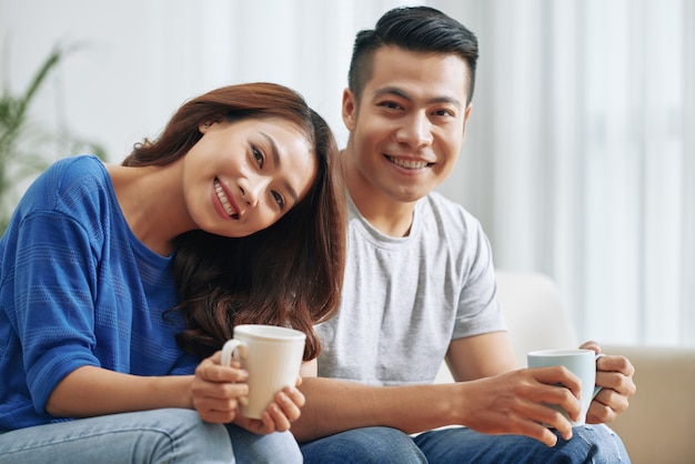 Happy Asian couple sitting on couch at home with tea mugs and smiling