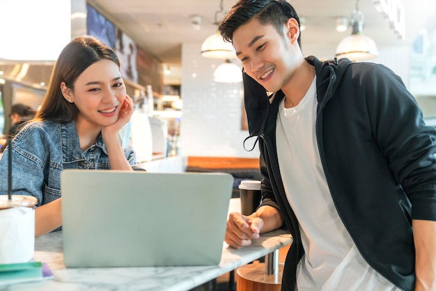 Happy asian couple take off face mask in a coffee shop surfing
internet on laptop young man and woman in a restaurant looking at
touch screen computer laugh smile together