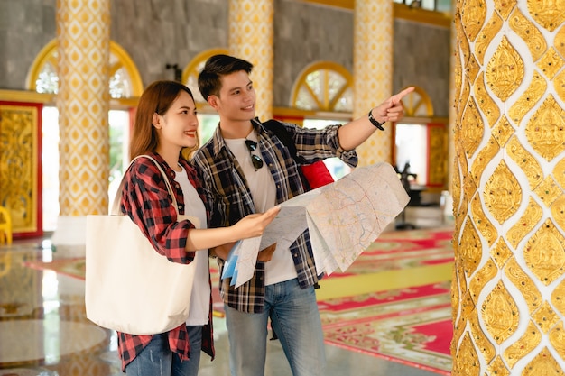 Счастливая азиатская пара туристических туристов, держащих бумажную карту и ищущих направление во время путешествия в тайский храм на отдыхе в Таиланде, красивый мужчина, указывая и проверяя карту