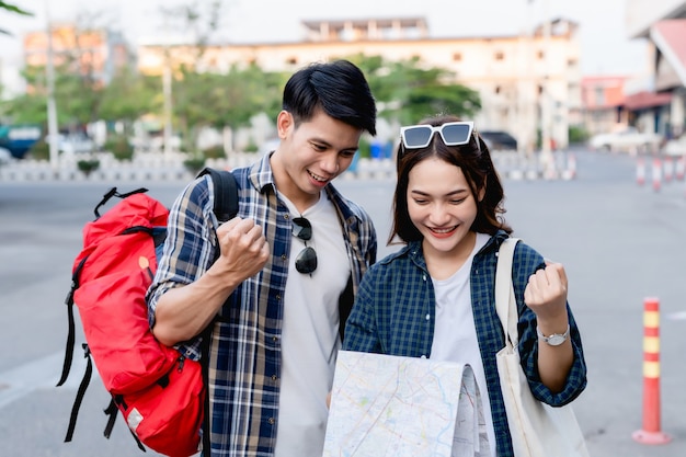 Happy Asian couple tourist backpackers holding paper map and looking for direction while traveling, They smile with glad when arrived at location on paper map destination.