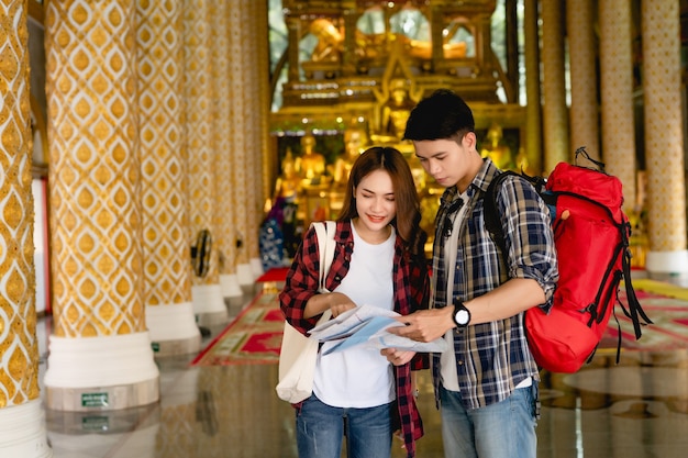 タイの休日に美しいタイの寺院を旅行しながら紙の地図で方向を探している幸せなアジアのカップルの観光バックパッカー