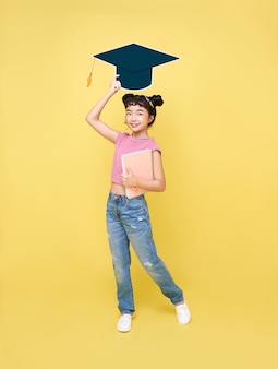Happy asian cute schoolgirl with graduation hat and book isolated on yellow background.