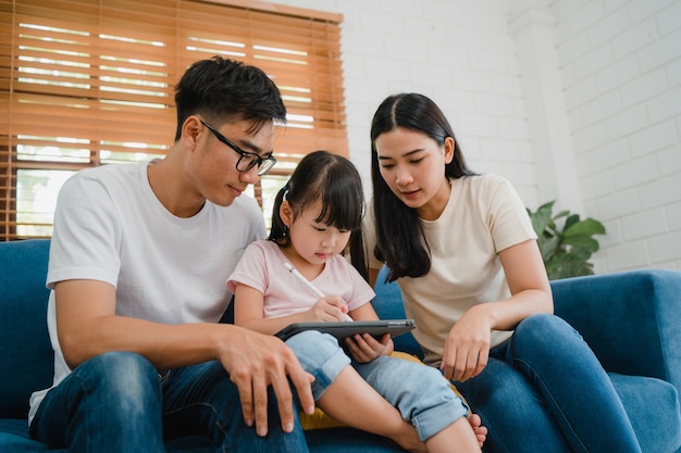 幸せなアジアの家族のお父さん、お母さんと娘が家のリビングルームでソファに座ってコンピュータータブレット技術を使用して