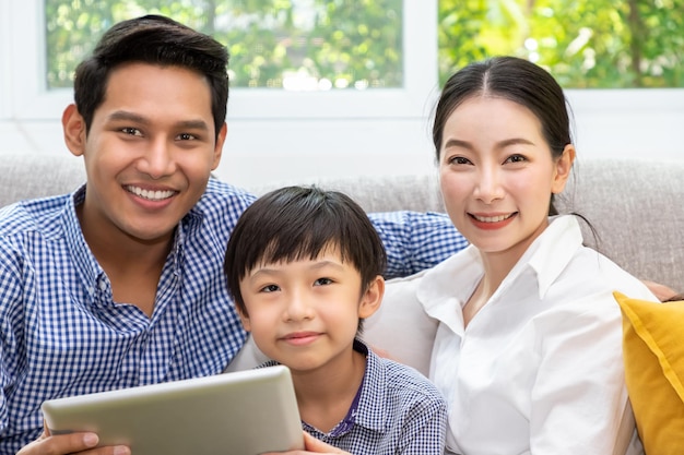 행복한 아시아 가족 아버지 어머니와 아들은 카메라를 보며 거실에서 태블릿을 함께 사용하여 소파에 앉아 있습니다.