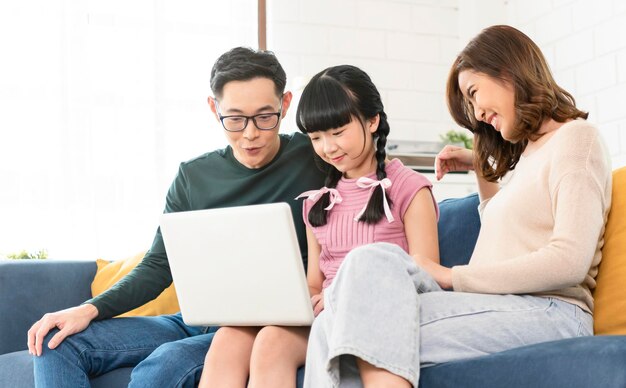 自宅のリビングルームxAのソファで一緒にコンピューターのラップトップを使用して幸せなアジアの家族