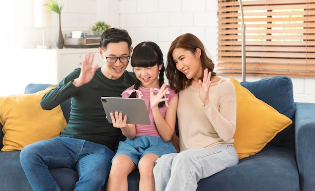 Happy Asian family using tablet video call virtual meeting together on sofa at home living room