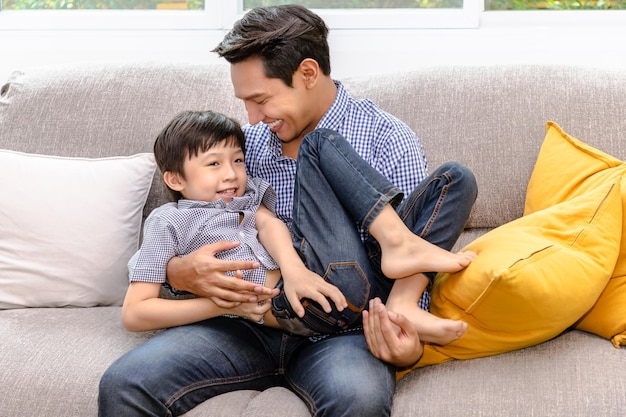 행복한 아시아 아버지는 가정의 가족 관계 개념에서 거실에 있는 소파에서 놀고 그의 아들을 안고 있습니다.