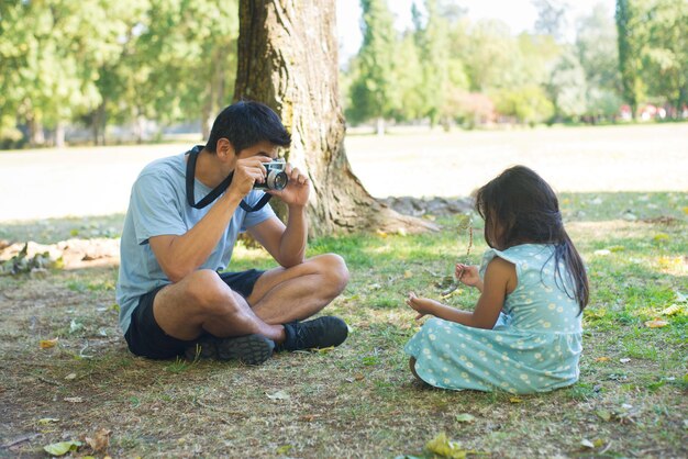 Happy Asian father taking photo of his daughter in park. Handsome man holding photo camera taking picture of little girl sitting on grassy field under trees. Leisure, hobby and happiness concept