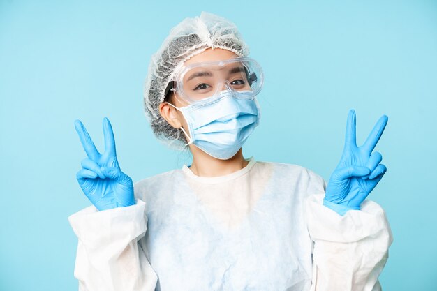 Счастливая азиатская женщина-врач, медсестра в средствах индивидуальной защиты, лицевой маске и стерильных перчатках, улыбается и показывает мир, v-знаки, синий фон.