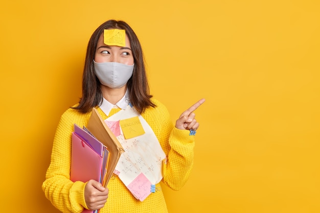 Счастливая азиатская женщина-менеджер, занятая оформлением документов, носит защитную одноразовую маску во время пандемии коронавируса и указывает указательным пальцем на место для копирования