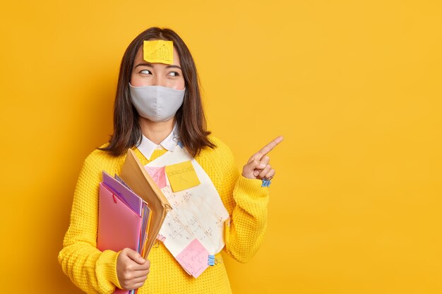 Happy Asian female manager busy doing paper work wears protective disposable mask during coronavirus pandemic indicates index finger on copy space