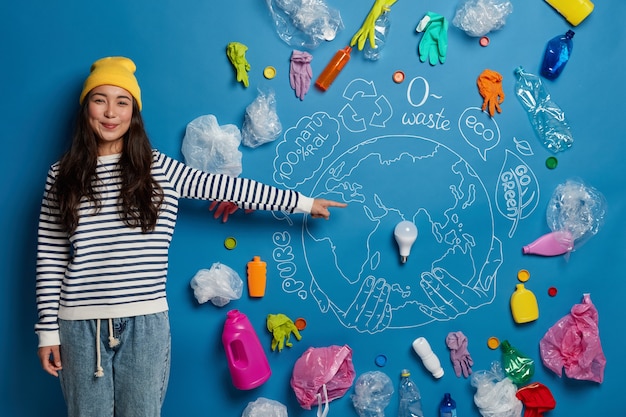 Happy Asian female volunteer prepares project about how to save Earth from pollution, demonstrates drawn planet with plastic garbage around