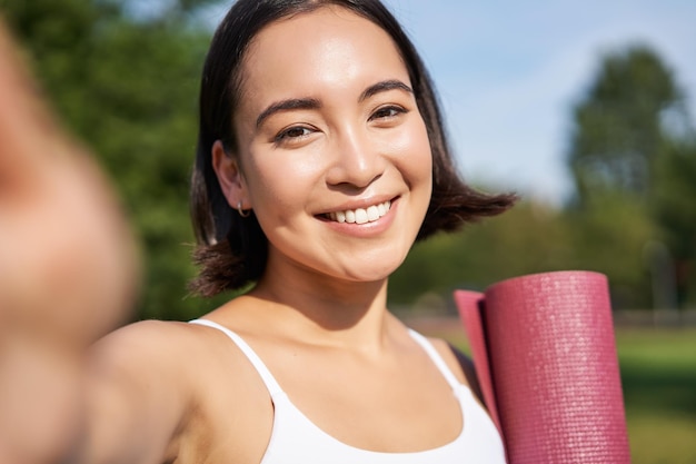 Free photo happy asian fitness girl takes selfie with rubber yoga mat in park healthy young sportswoman makes p