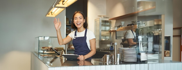 Happy asian girl barista say next please raise arm to invite cafe clients to order coffee standing