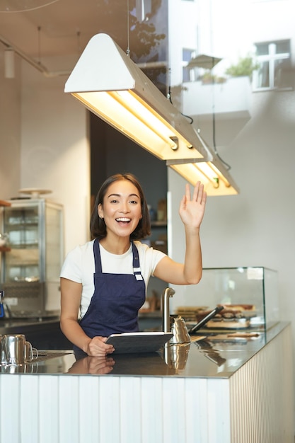 Happy asian girl barista works in coffee shop waves at client processing orders behind counter in ca