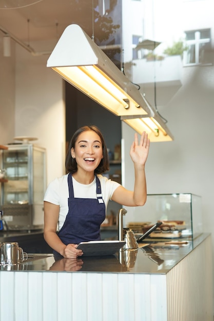 幸せなアジアの女の子のバリスタは、カリフォルニア州のカウンターの後ろで注文を処理するクライアントで喫茶店の波で働いています