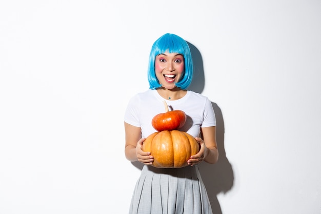 Happy asian girl in blue wig holding two cute pumpkins and smiling at camera, wearing schoolgirl outfit for halloween party.