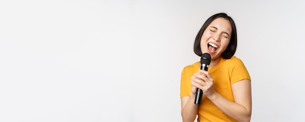 Happy asian girl dancing and singing karaoke holding microphone in hand having fun standing over whi