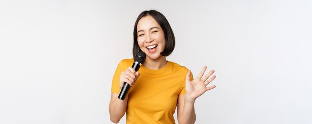 Happy asian girl dancing and singing karaoke holding microphone in hand having fun standing over whi