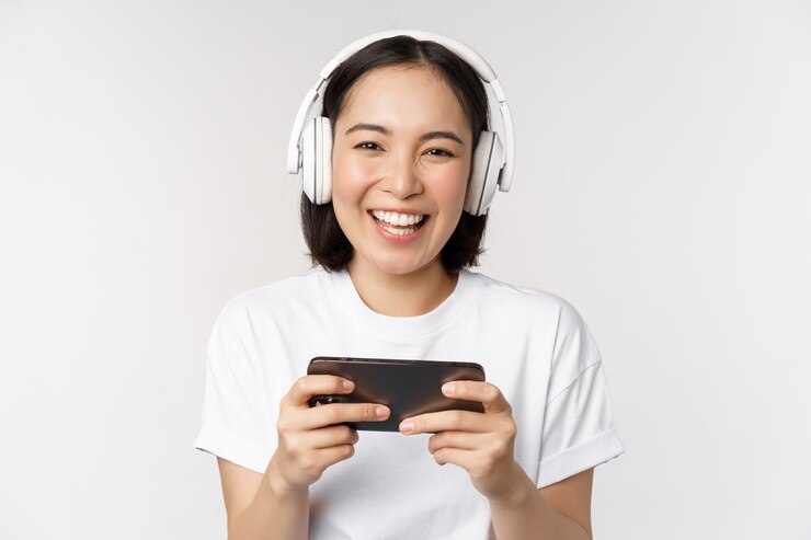 無料Photo 白い背景の上に立っているヘッドフォンを身に着けているスマートフォンで見ている携帯電話で遊んで幸せなアジアの女の子ゲーマー